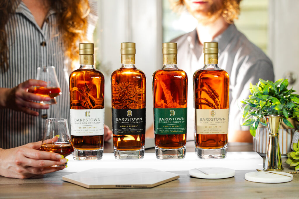 Four Bardstown Bourbon Company Origin Series bottles with amber bourbon, held and tasted by two people.