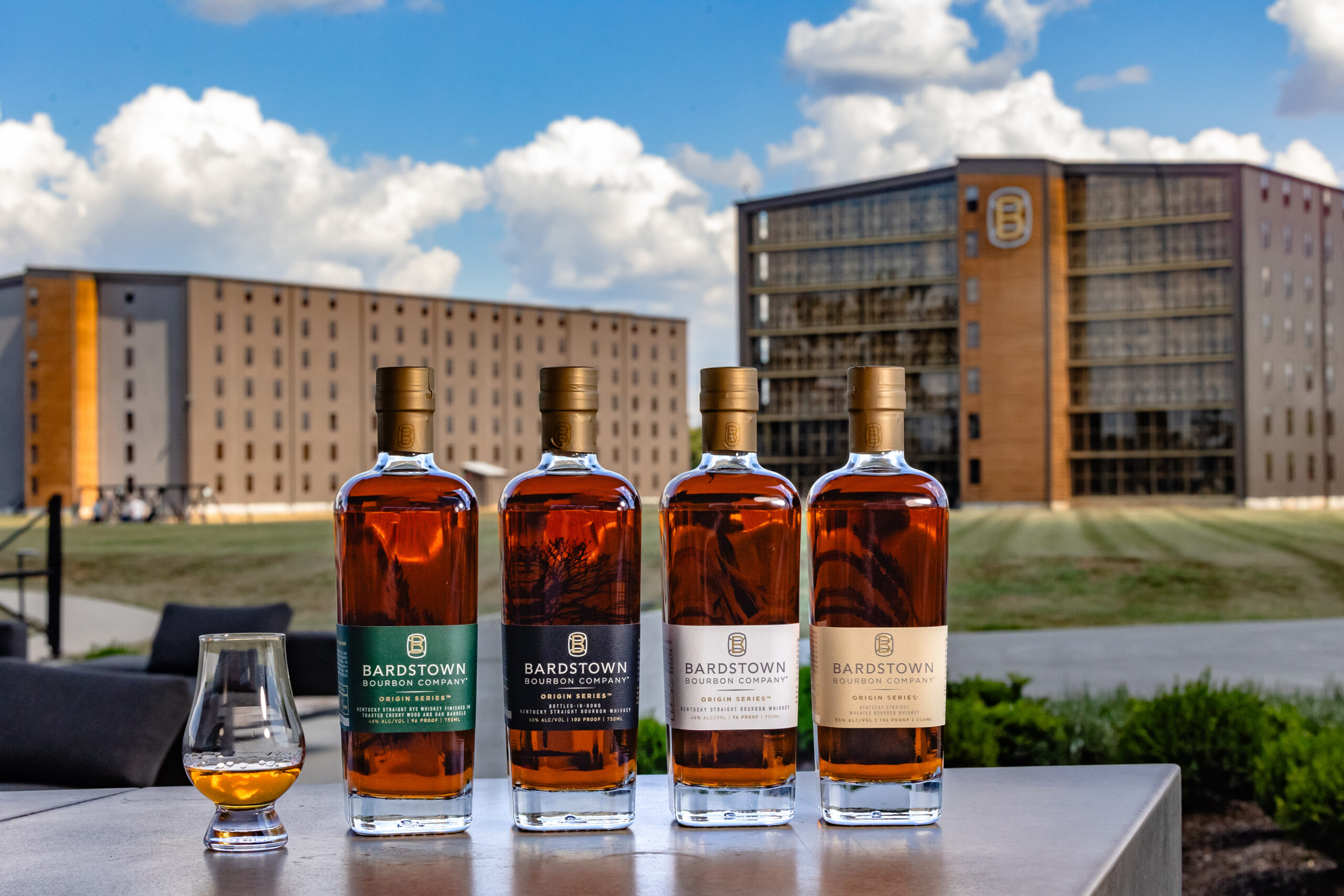 Four Bardstown Bourbon bottles lined up with a glass pour, distillery buildings in the background under the blue sky.