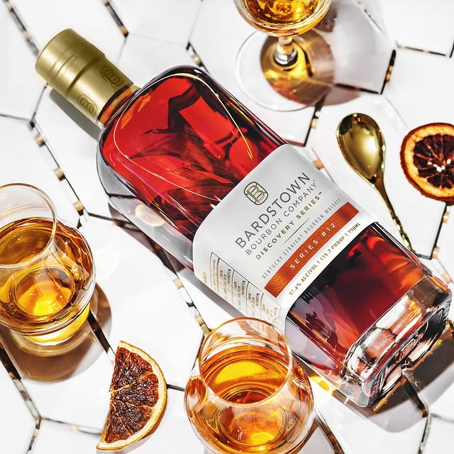 Bardstown Bourbon Company Discovery Series bottle with poured glasses, dried orange slices, and a gold spoon on a white tile surface.